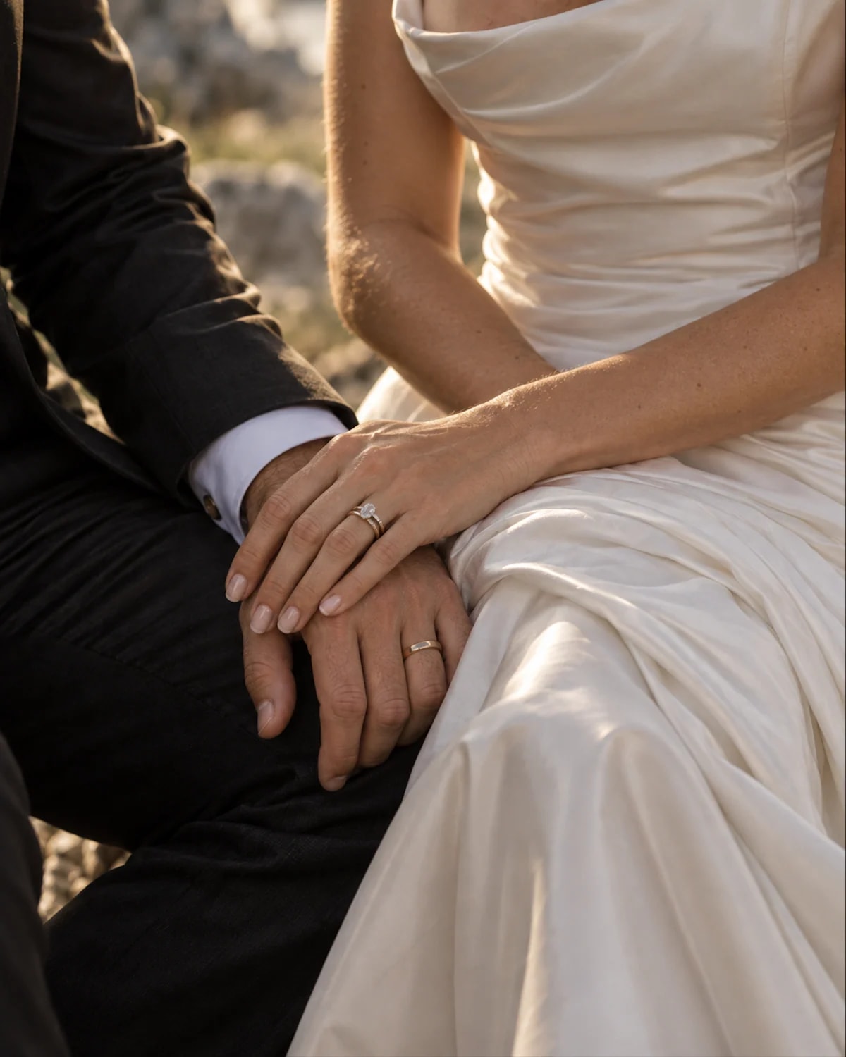 A bride and groom resting their hands together, showing their wedding rings in the evening light.