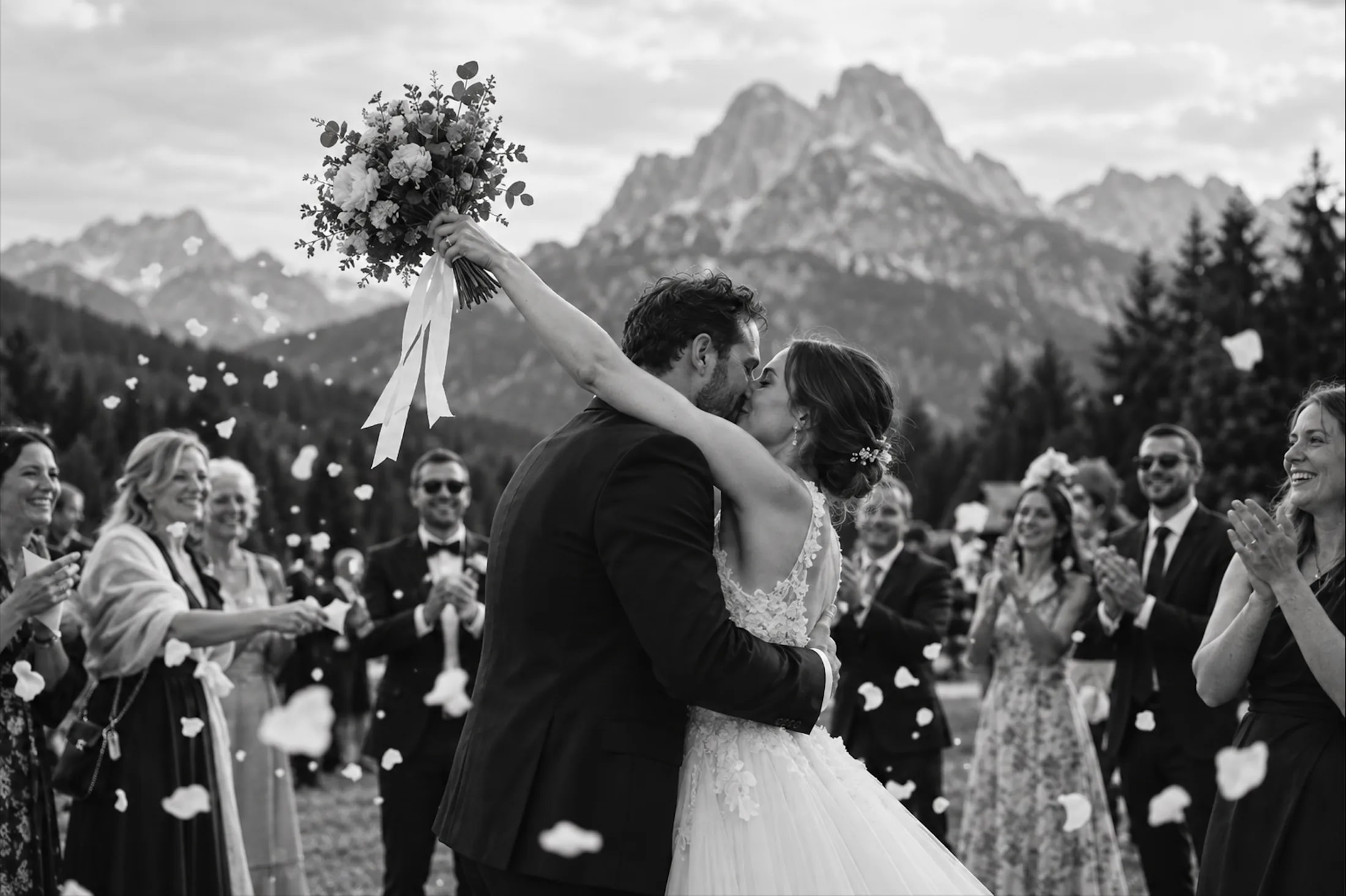 Newlyweds kiss with wedding guests celebrating against a Dolomites mountain backdrop.