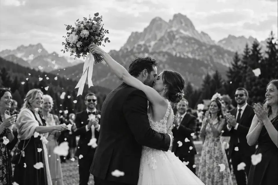 Newlyweds kiss with wedding guests celebrating against a Dolomites mountain backdrop.
