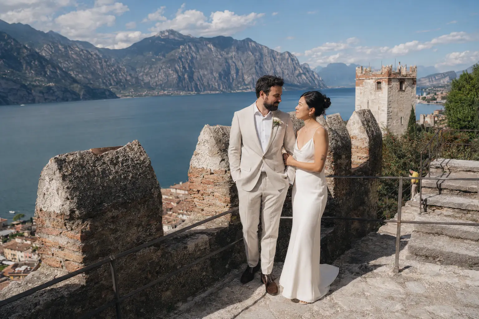 Alt text: Bride and groom standing on the medieval walls of Castello Scaligero di Malcesine overlooking Lake Garda and the surrounding mountains during a.