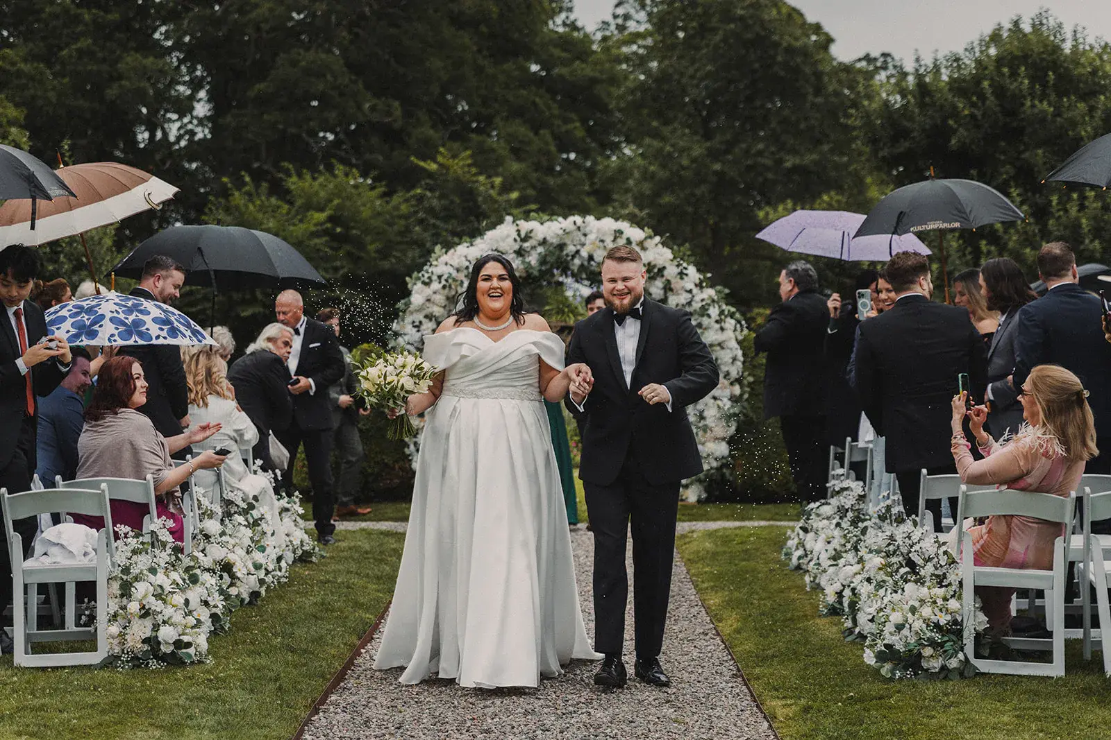 Newlyweds walking down the garden aisle among guests with umbrellas at Södertuna Slott.